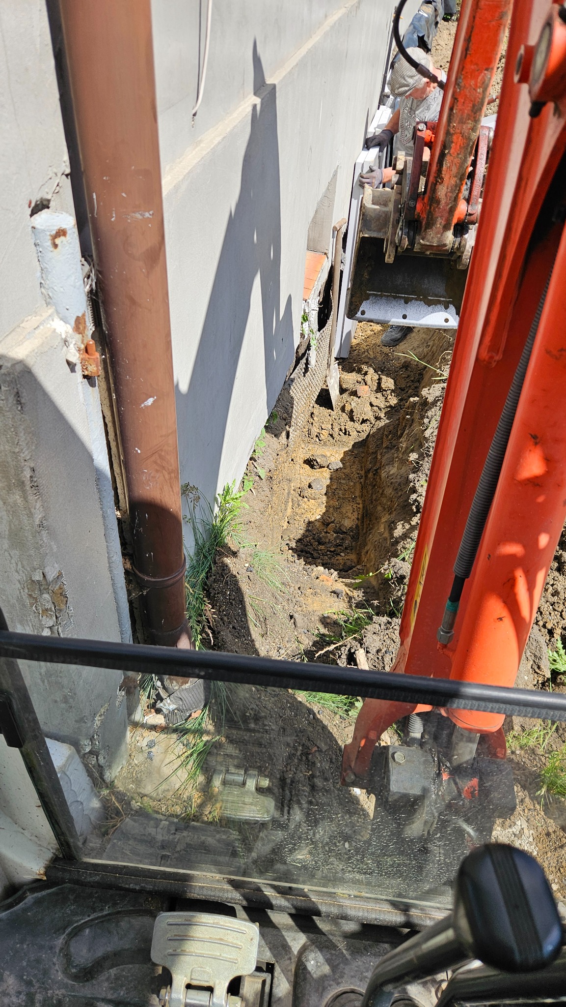 Wykop wąskiego rowu obok budynku, wykonany pomarańczową mini koparką. Widok z kabiny operatora, z bliska widoczny rów i fragment ściany budynku.
