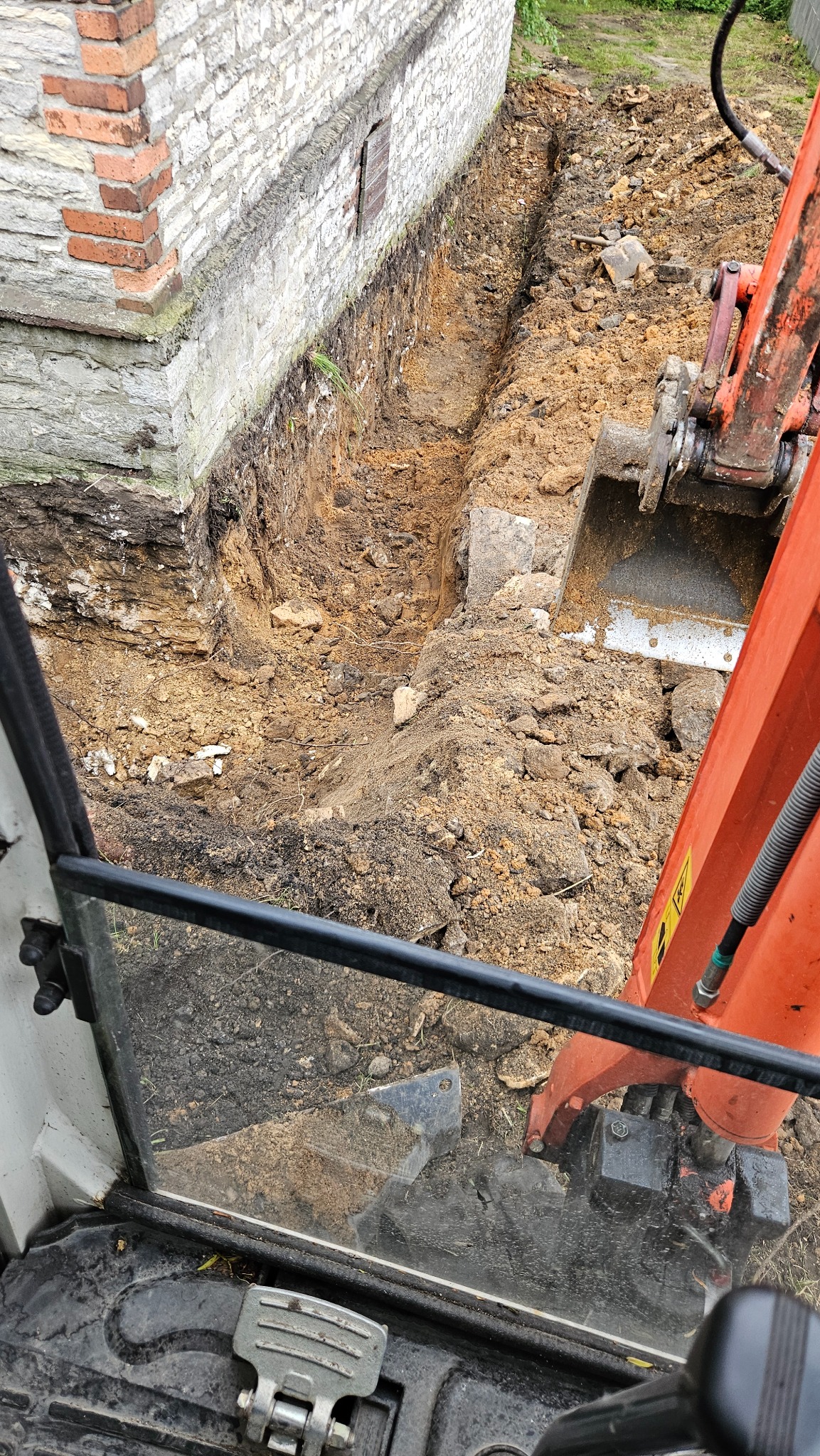 Wąski wykop w ziemi, widoczny z kabiny koparki obok murowanej ściany z ceglanymi detalami. Pomarańczowe ramię koparki częściowo zasłania widok.
