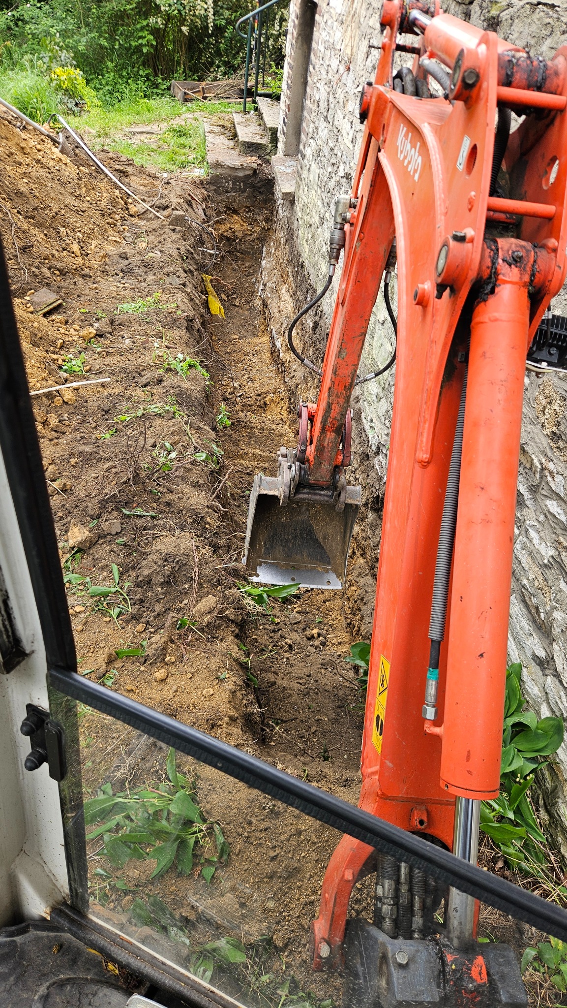 Pomarańczowa koparka Kubota w trakcie wykopu wąskiego rowu wzdłuż kamiennej ściany budynku. Widok z kabiny operatora, częściowo widoczna szyba.