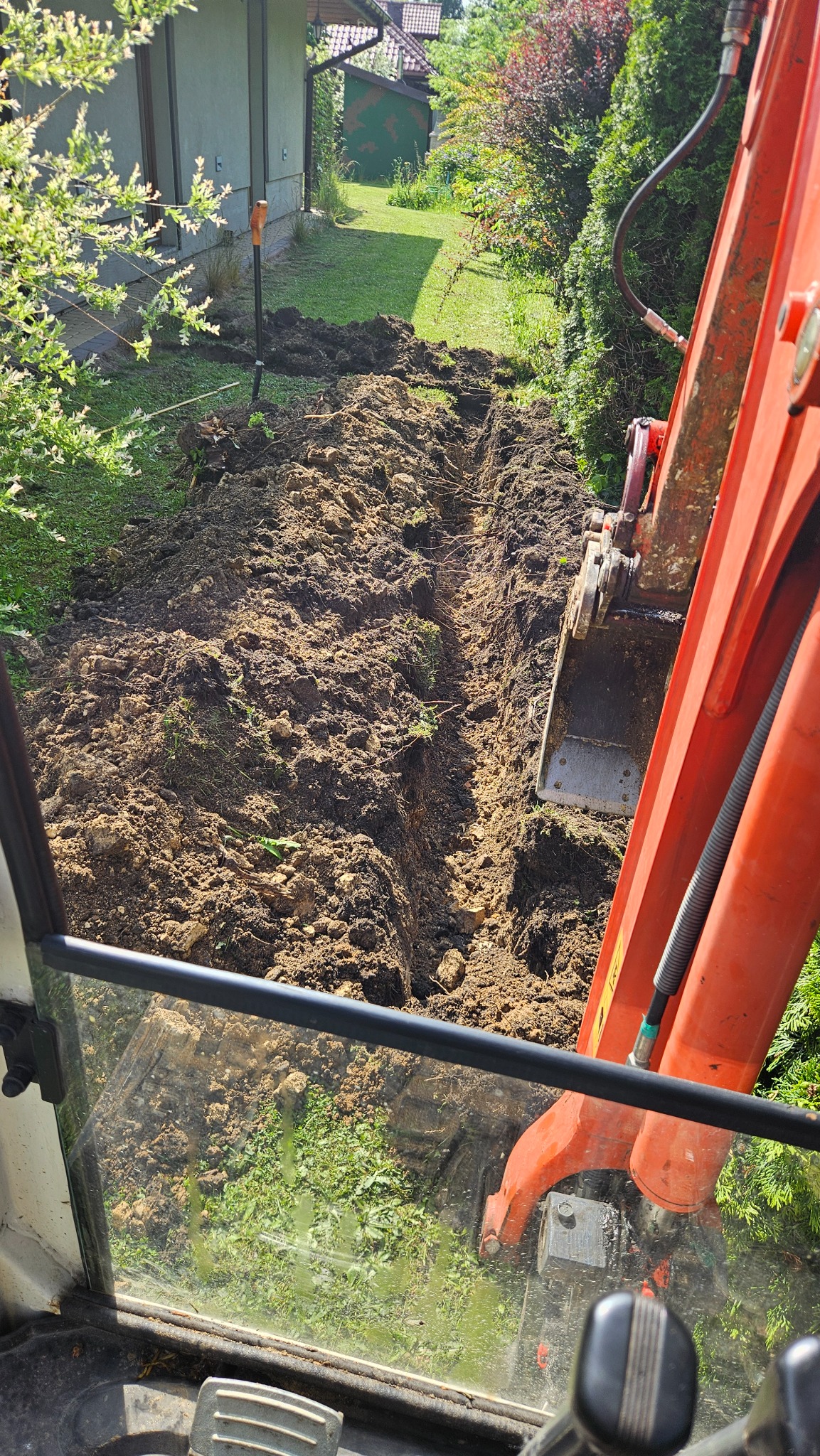 Wykop wąskoprzestrzenny pod instalacje w Jaworznie, widziany z kabiny koparki. W tle zielony trawnik i budynek. Ślady pracy łopaty.