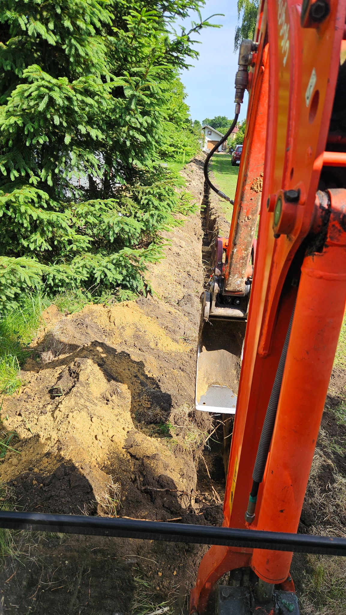 Pomarańczowa koparka w trakcie wykonywania wąskiego wykopu w ziemi, tuż obok zielonych drzew iglastych w słoneczny dzień. Widok z perspektywy operatora.