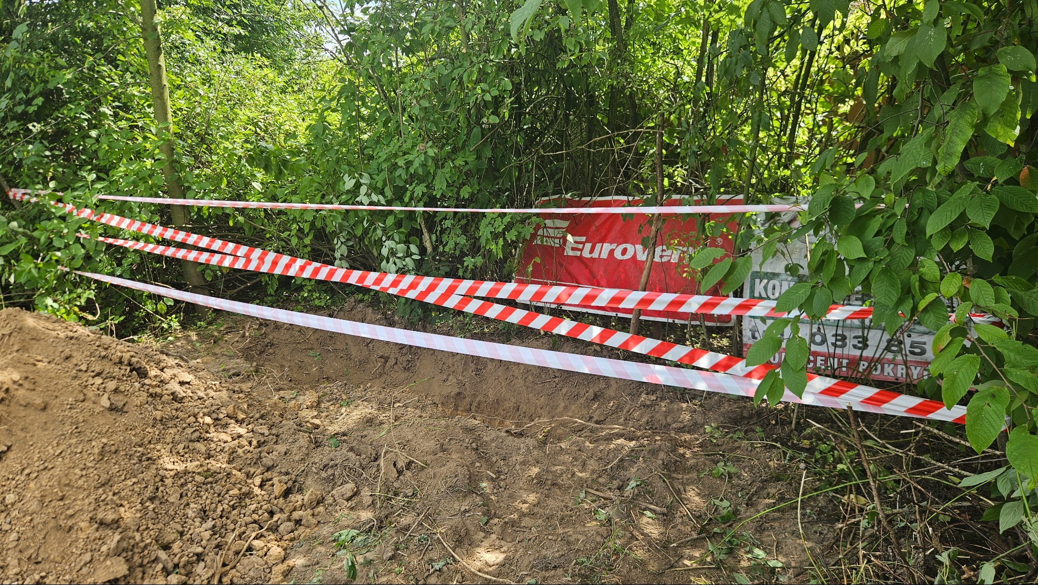 Wykop ziemny zabezpieczony taśmą ostrzegawczą w biało-czerwone pasy, w tle zieleń i tablica reklamowa. Ujęcie z poziomu gruntu.