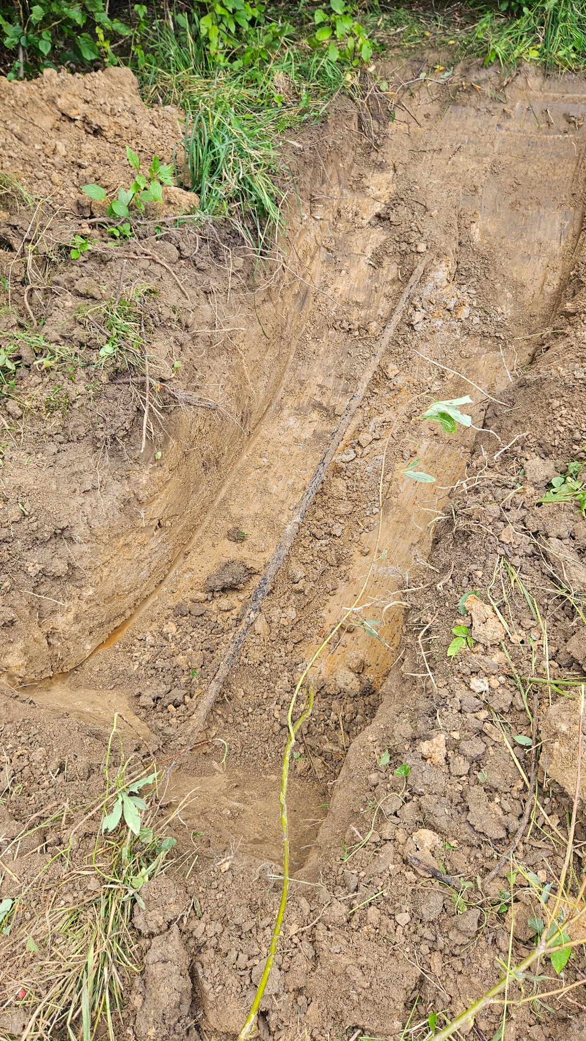 Wąski wykop w ziemi z widoczną rurą. Ściany wykopu odsłaniają warstwy gleby. Ziemia wokół wykopu porośnięta trawą i roślinnością.