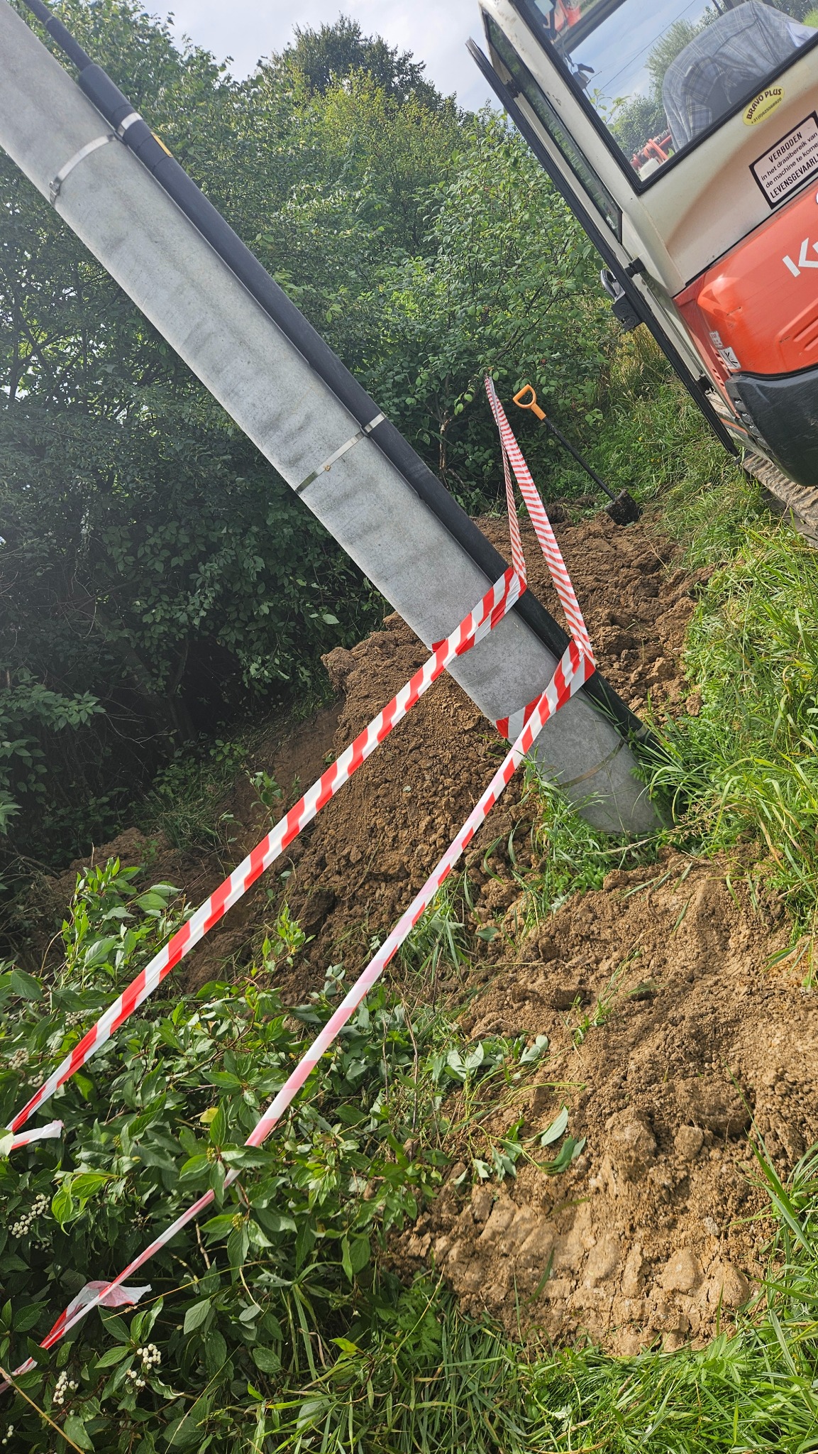 Skośne ujęcie rury drenażowej z czarną osłoną, zabezpieczonej taśmą ostrzegawczą na zboczu ziemnym, z widocznym fragmentem koparki i zielenią w tle.