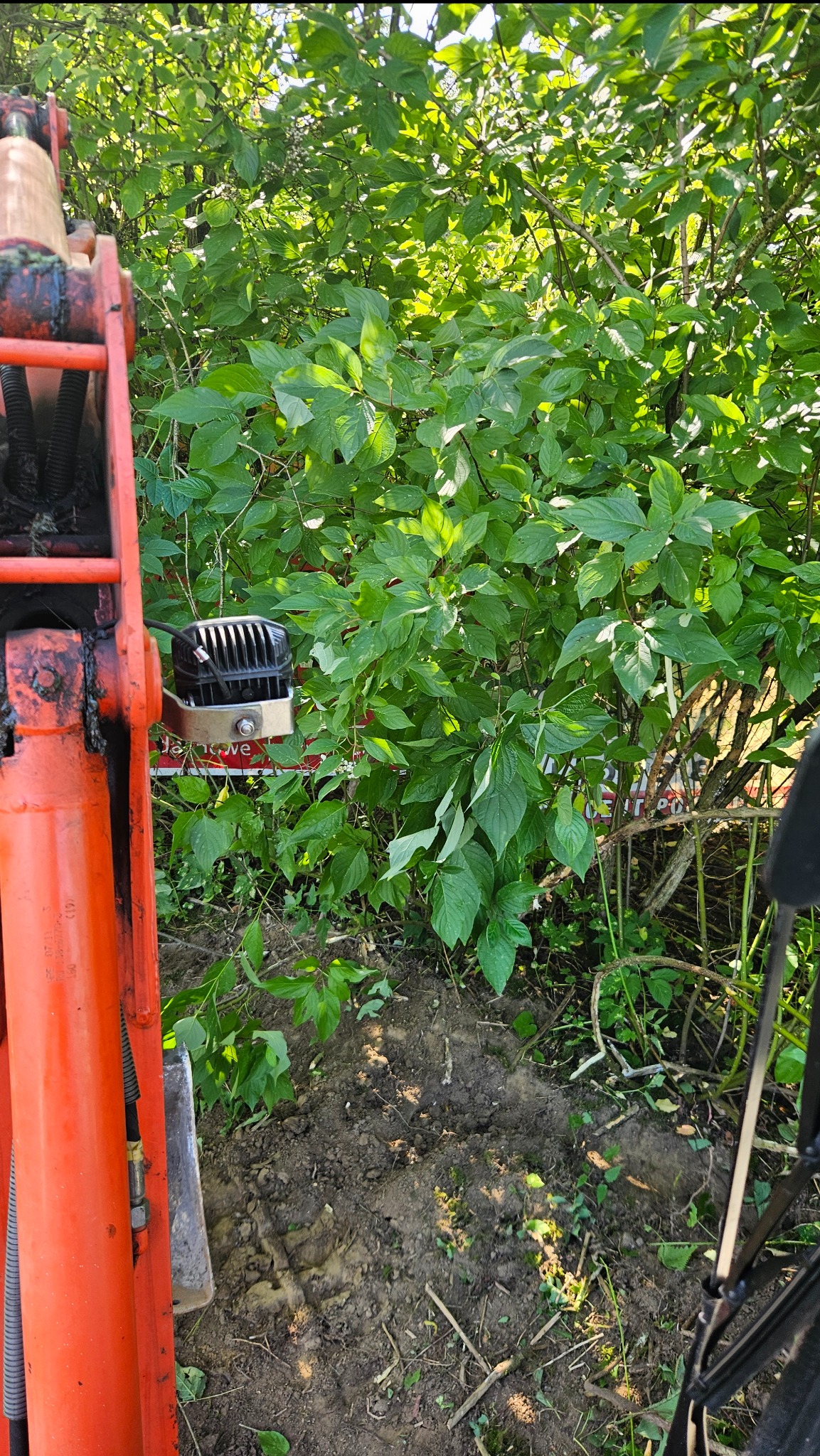 Fragment pomarańczowego ramienia koparki w trakcie pracy przy zaroślach i wykopanym gruncie, z widocznym oświetleniem roboczym.