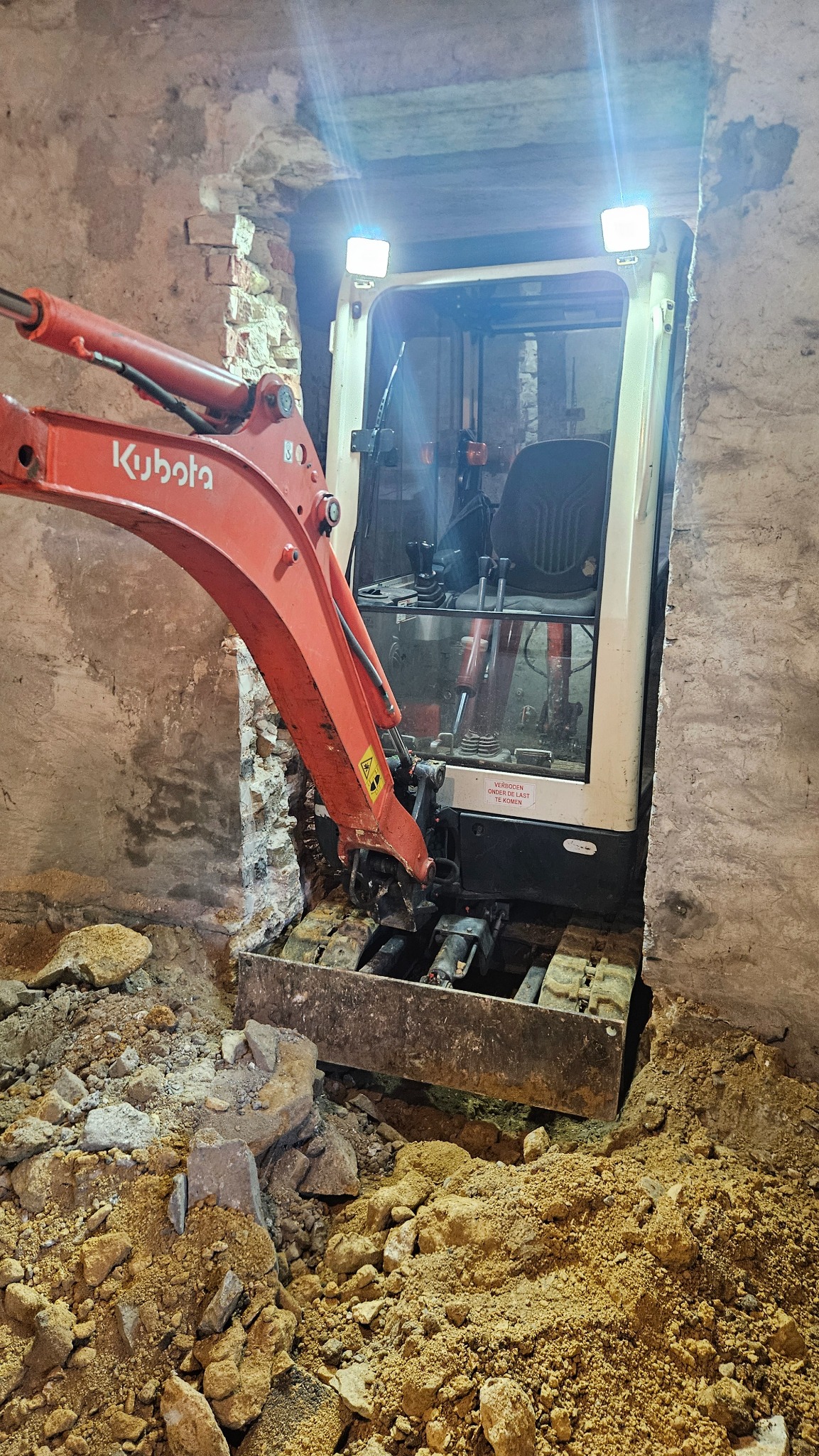 Pomarańczowa mini koparka Kubota w wąskim wykopie wewnątrz budynku z kamiennymi ścianami. Oświetlenie koparki skierowane na wykop.