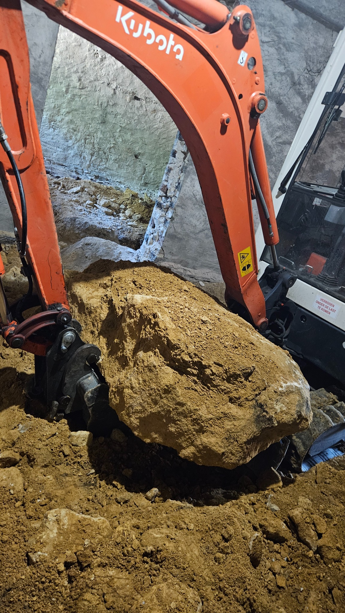 Pomarańczowa koparka Kubota w trakcie podnoszenia dużego, beżowego kamienia/skały w wykopie. Widoczny operator koparki i fragment ściany wykopu z elementami cegieł.