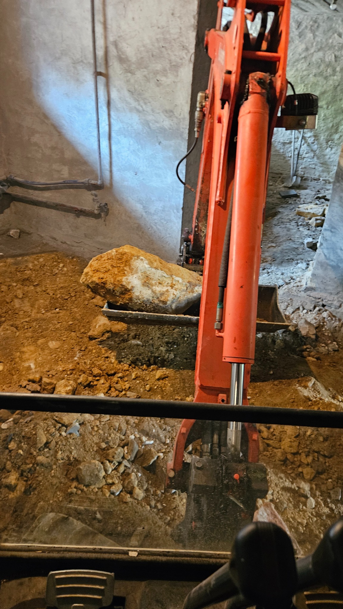 Pomarańczowa koparka w wykopie, usuwa duży kamień. Widok z kabiny operatora, ściany wykopu oświetlone, widoczne rury na ścianie.
