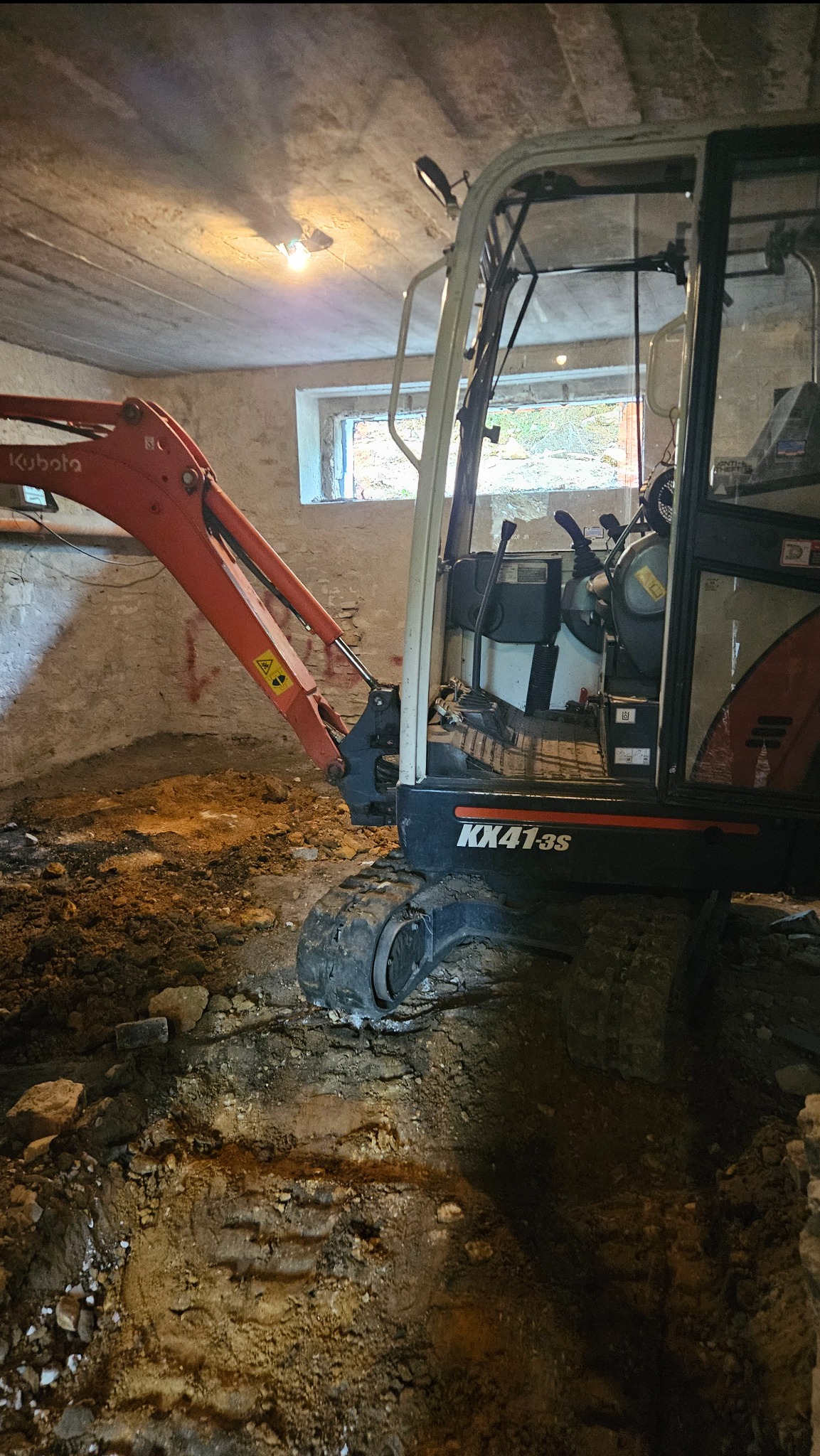 Mini koparka Kubota KX41-3S w trakcie prac ziemnych wewnątrz budynku, widoczny wykop i fragment okna. Oświetlenie sztuczne.