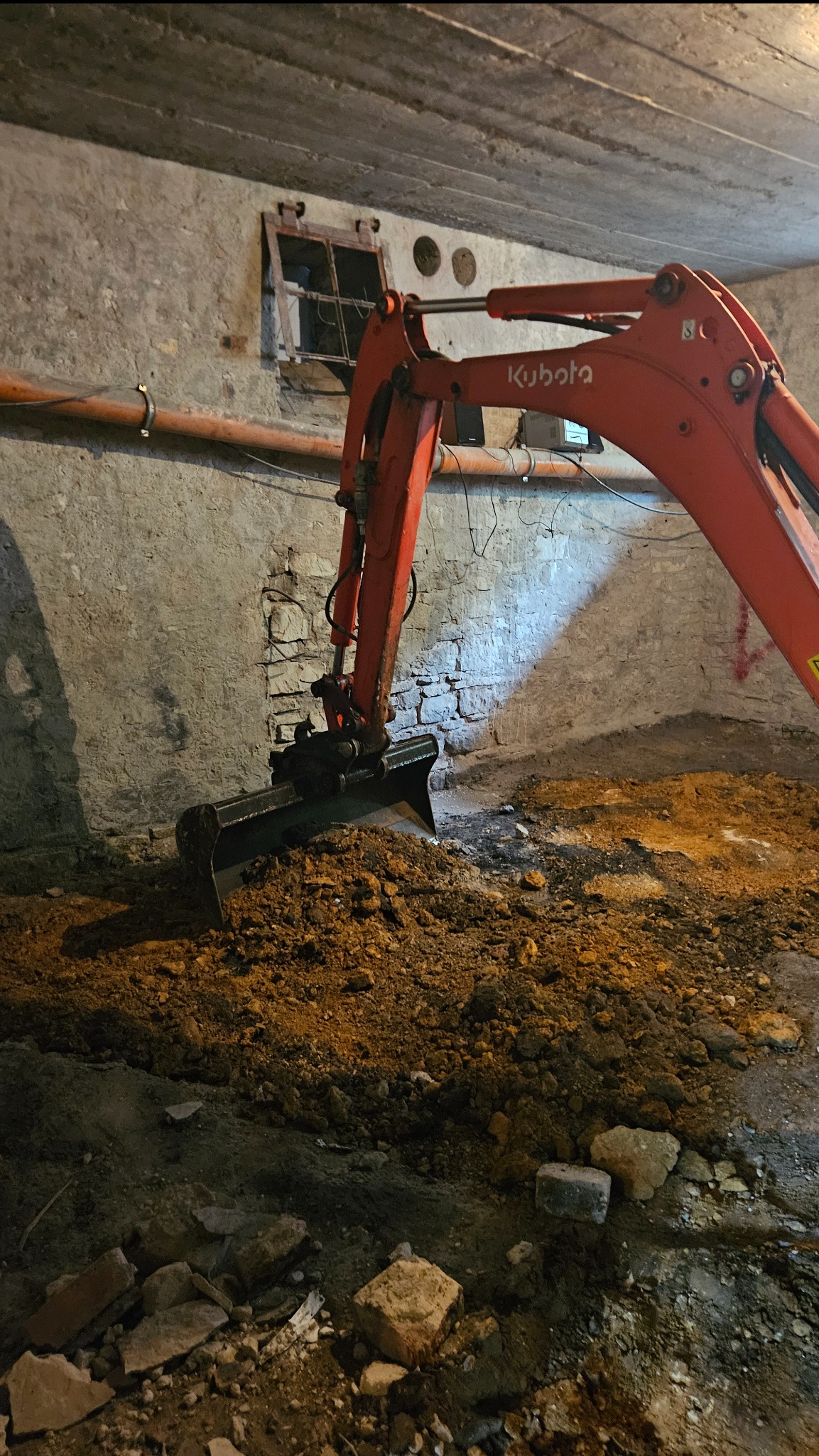 Pomarańczowa mini koparka Kubota w trakcie prac ziemnych w piwnicy oświetlonej światłem, widoczne ściany z cegły i betonowy strop.