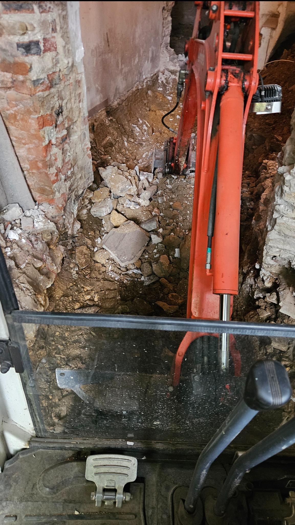Wąski wykop w ziemi, widok z kabiny koparki z dźwigniami sterującymi, ściany wykopu z cegły i kamienia, pomarańczowe ramię koparki wewnątrz wykopu.