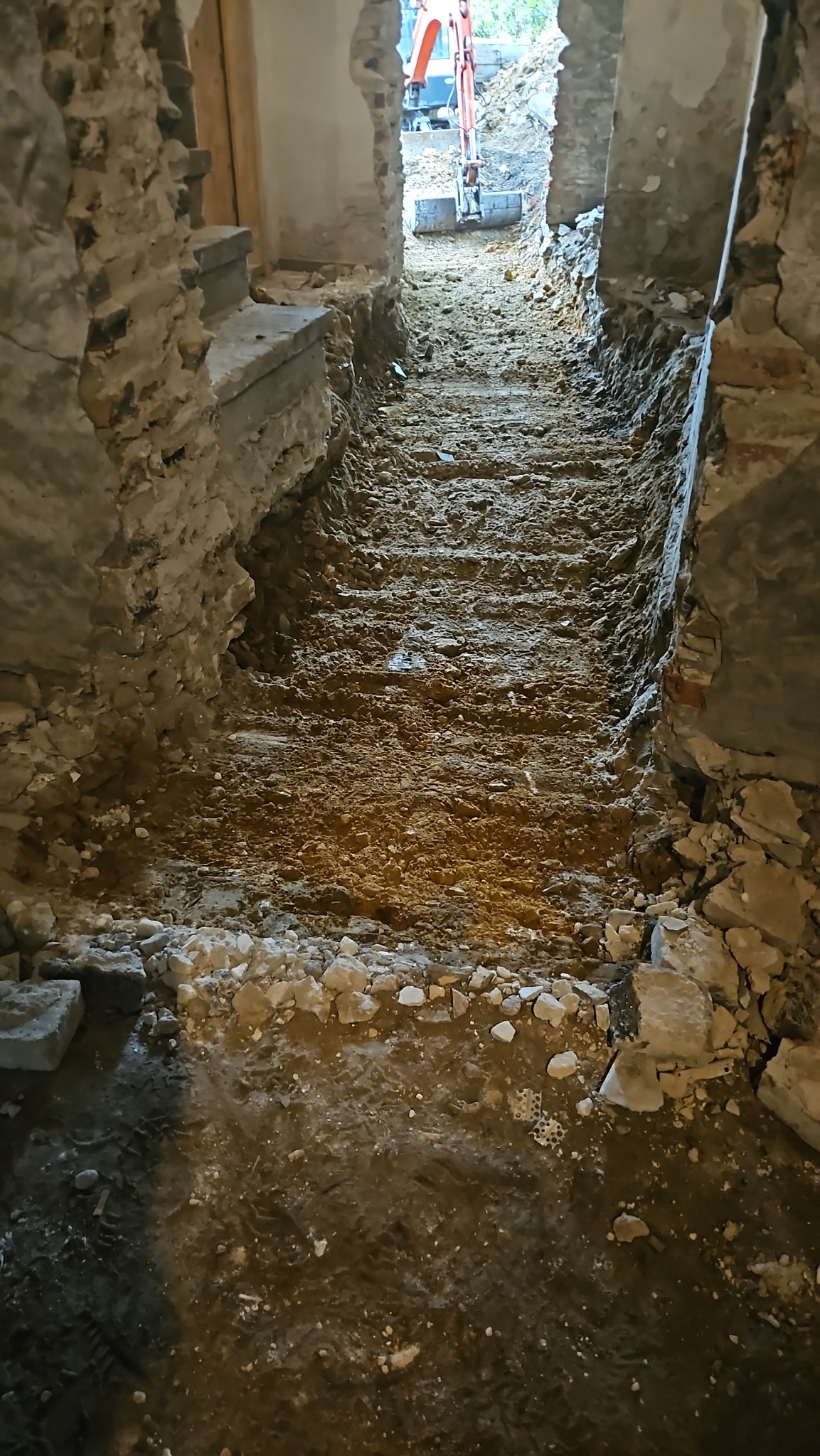 Wąski wykop wewnątrz budynku z odsłoniętymi fundamentami i mini koparką w tle. Widoczne ślady opon i gruz.