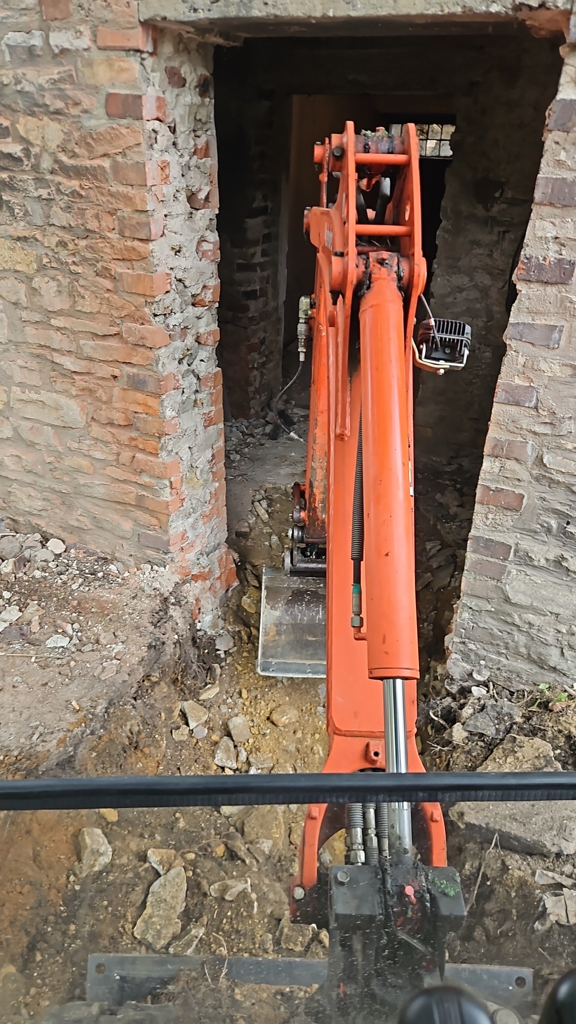 Pomarańczowa mini koparka w trakcie prac ziemnych, widoczna z perspektywy operatora, w tle ceglany mur z otworem.