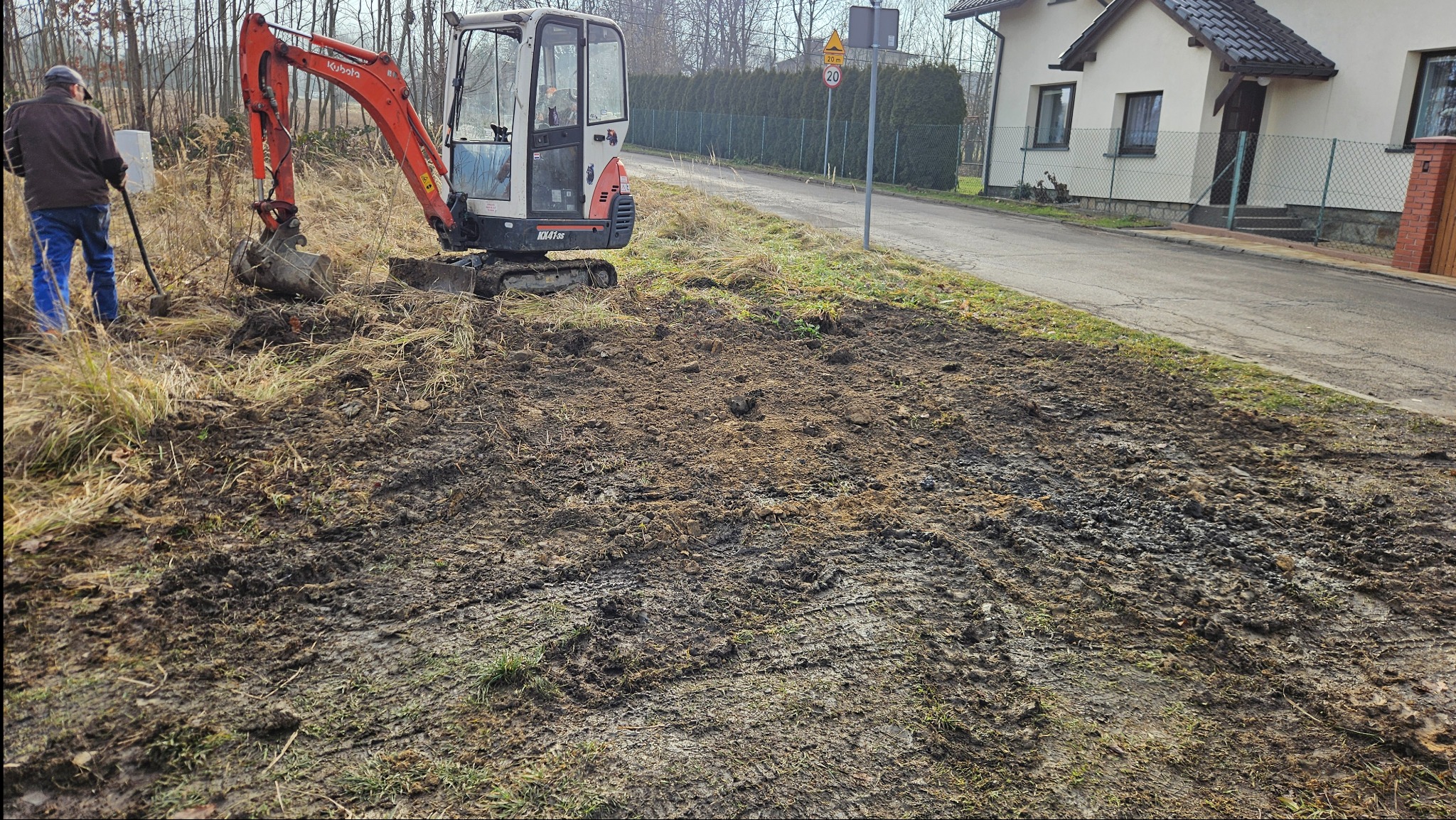 Pomarańczowa mini koparka Kubota w trakcie niwelacji terenu przy drodze i budynku. Operator koparki stoi obok z łopatą. Widoczne ślady opon i wykopana ziemia.