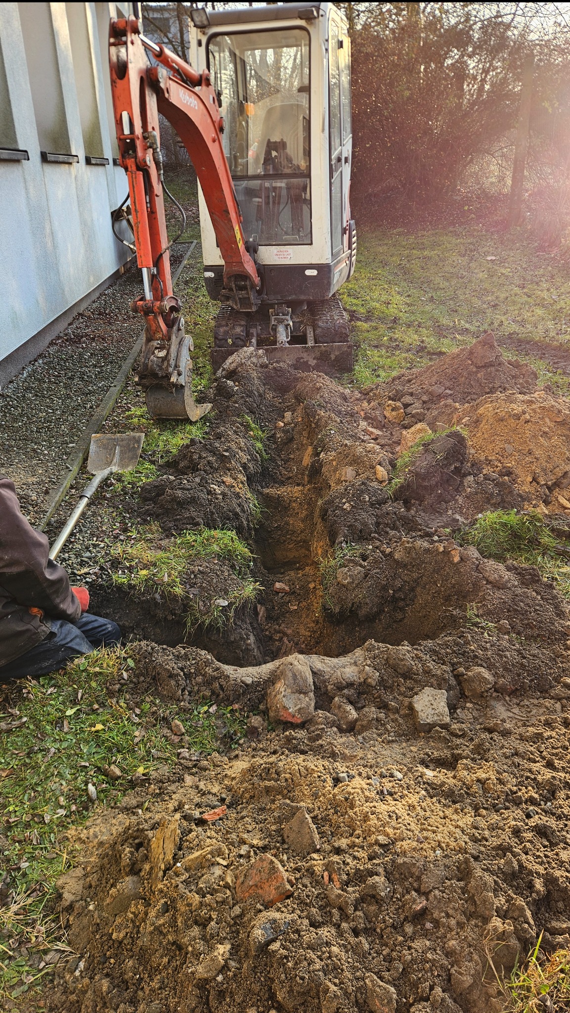 Mini koparka Kubota przy wykopie wąskiego rowu w ziemi z zieloną trawą, obok leży łopata i widać fragment osoby w roboczym ubraniu.