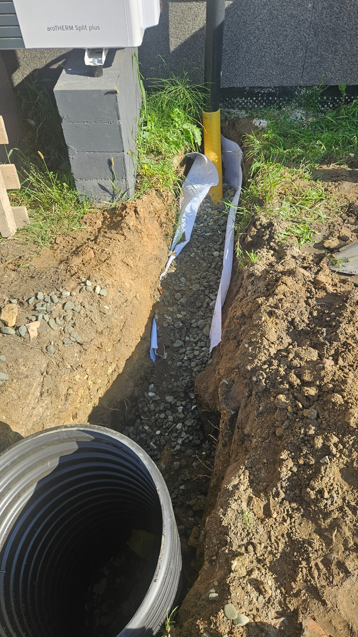 Wykopany rów z żółtą rurą drenażową, obsypany kamieniami i zabezpieczony białą geowłókniną. W rogu widoczny czarny zbiornik. Prace ziemne w ogrodzie.