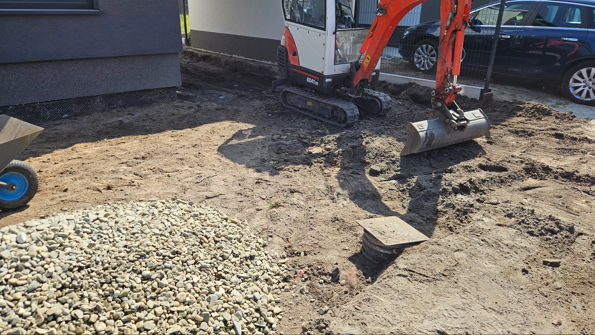 Mini koparka na placu budowy obok domu, z kupką kamieni i wózkiem. Widoczny fragment fundamentu. Prace ziemne w słoneczny dzień.