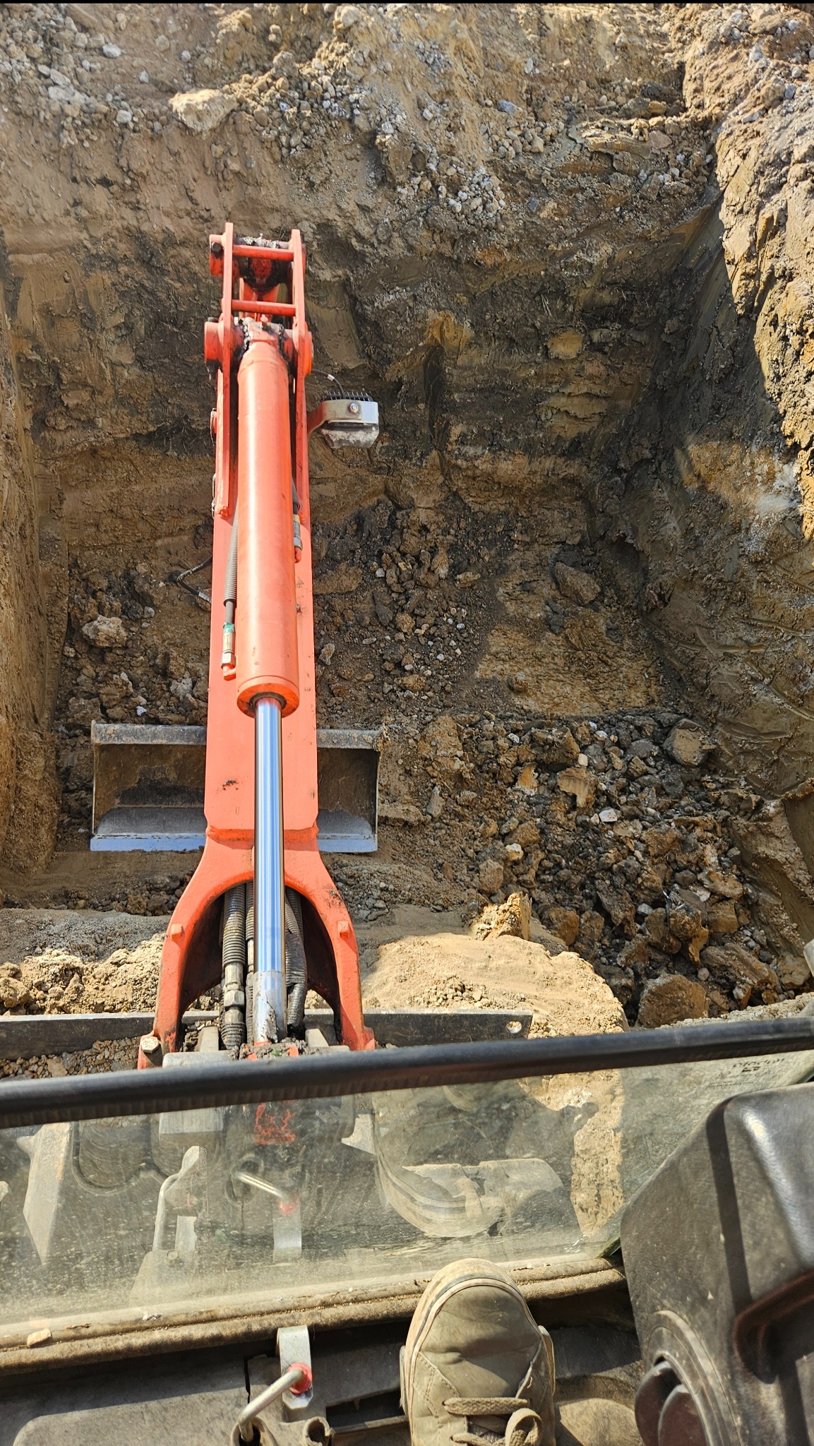 Pomarańczowa ramię koparki w głębokim wykopie ziemnym, widok z kabiny operatora, z fragmentem buta w kadrze. Wykop o nierównych ścianach, z widoczną strukturą gleby.