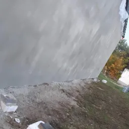 Świeżo nałożona, jasnoszara warstwa tynku na zewnętrznej ścianie budynku, widoczna z bliskiej perspektywy, z fragmentami gruzu i ziemi u podstawy ściany, sugerującymi trwające prace wykończeniowe.