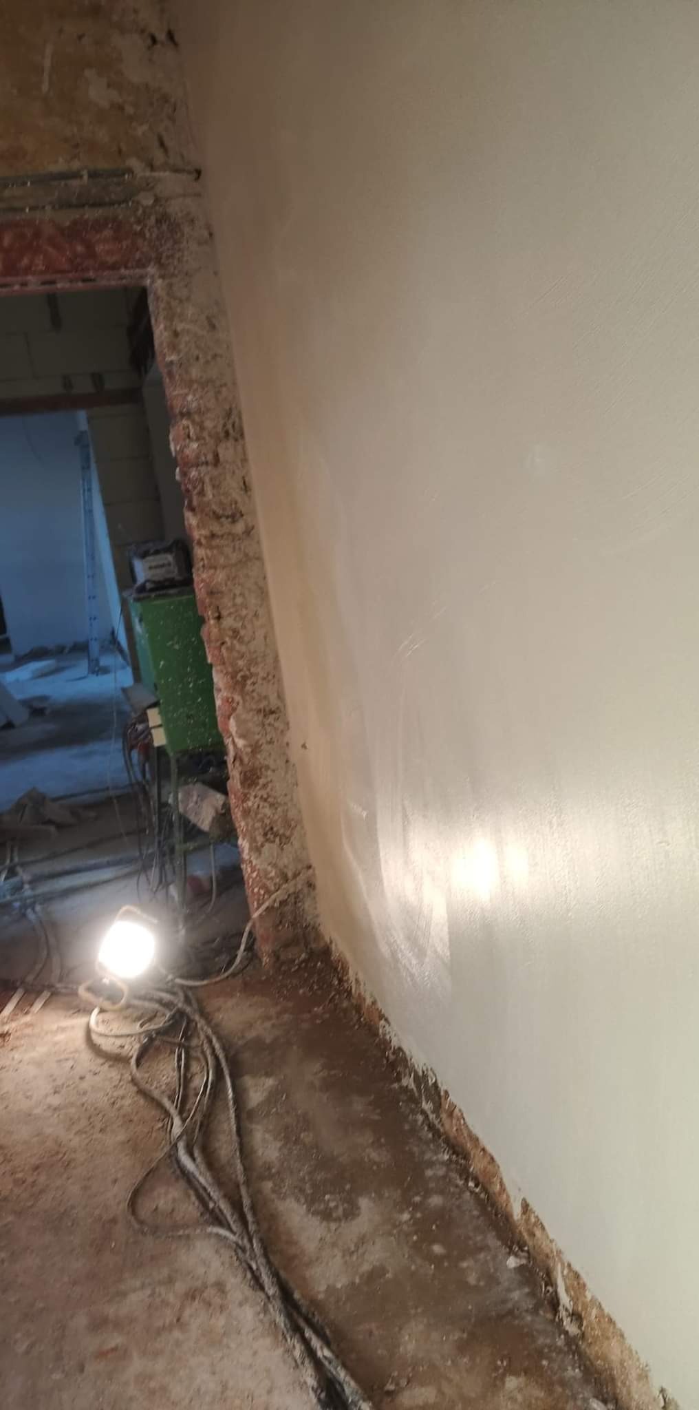 Świeżo wyszpachlowana, gładka ściana w trakcie remontu, widoczne kable oświetleniowe na podłodze i surowy mur w przejściu do innego pomieszczenia.