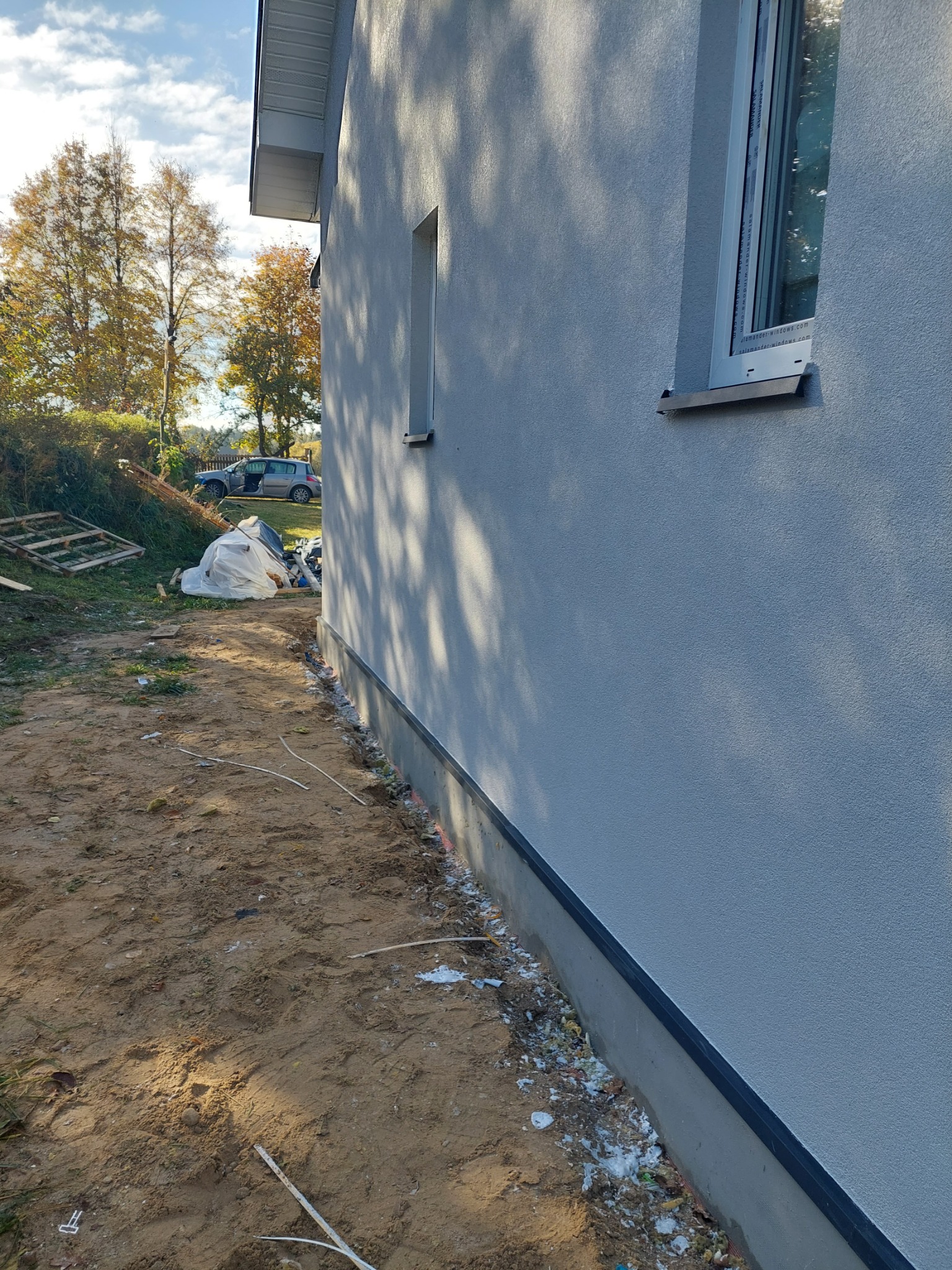 Świeżo otynkowana ściana budynku w odcieniach szarości, widoczne okno z białymi ramami, teren wokół w trakcie prac porządkowych, w tle drzewa i zaparkowany samochód z otwartymi drzwiami.