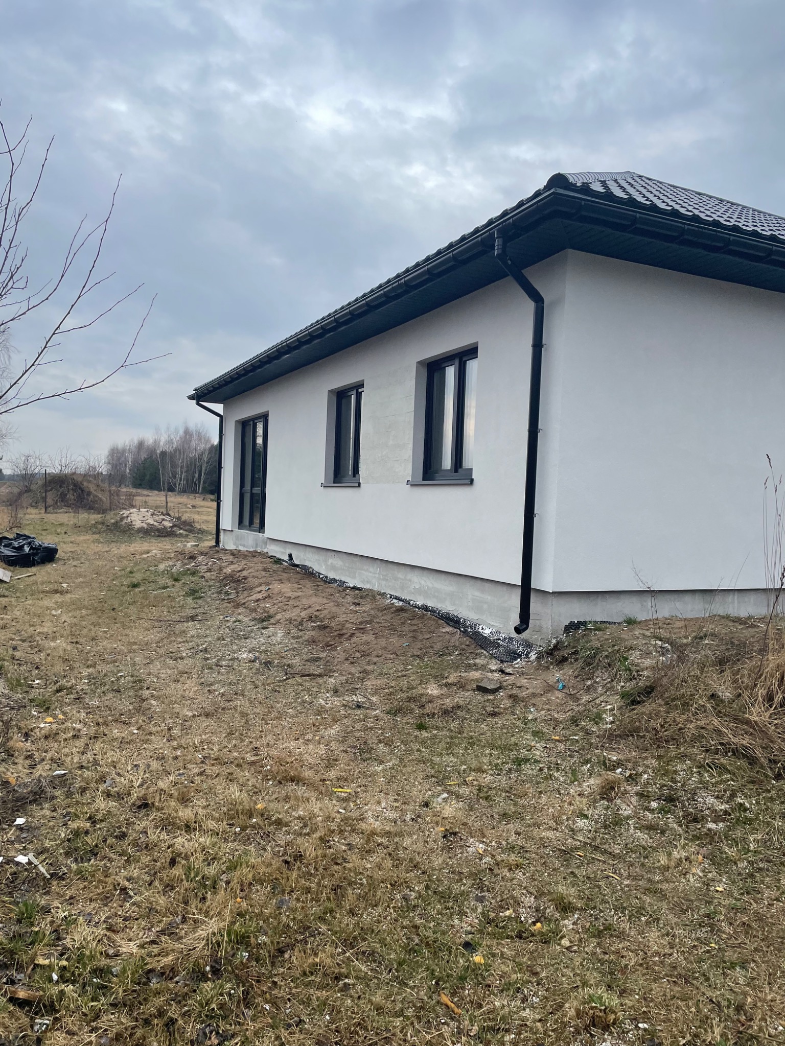 Wykończona elewacja domu jednorodzinnego z czarnymi oknami i rynnami, widok z boku na tle pochmurnego nieba i niezagospodarowanego terenu.