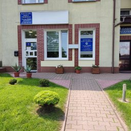 Kancelaria Radcy Prawnego Radca Prawny Joanna Wołek - Wejście do budynku z tabliczkami informacyjnymi o Kancelarii Radcy Prawnego Joanna Wołek i Gabinecie Fizjoterapii, z zadbanym trawnikiem, krzewami i kwiatami w donicach.