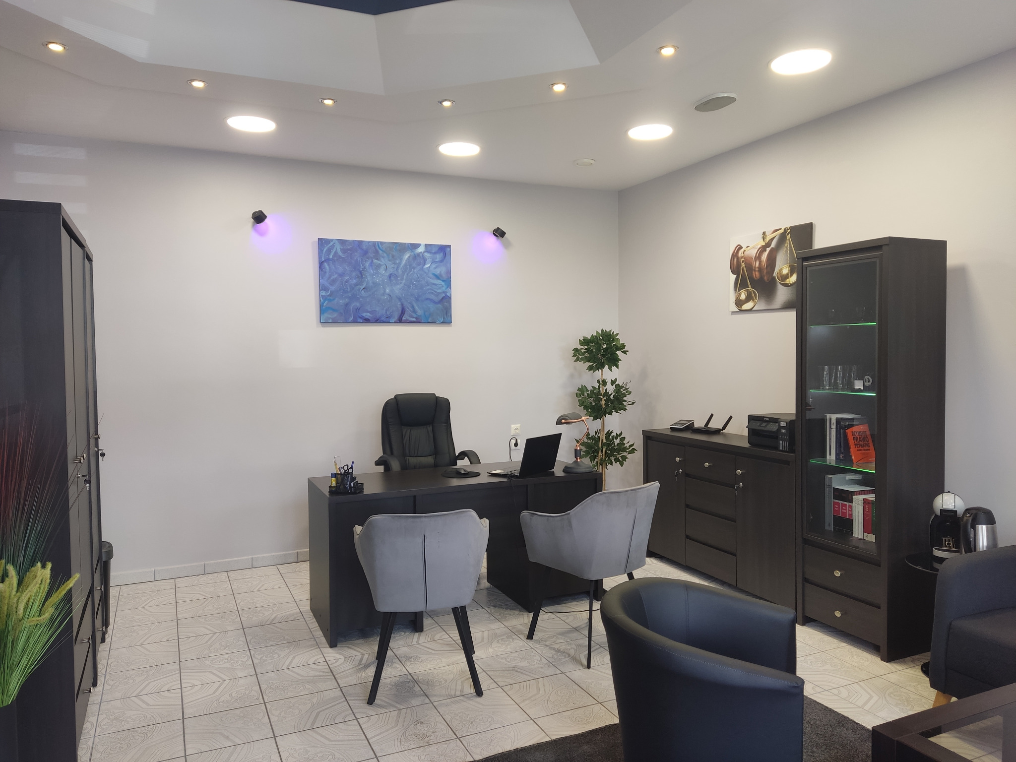 Nowoczesne biuro prawnicze z ciemnymi meblami, szarymi krzesłami i abstrakcyjnym obrazem na ścianie, oświetlone punktowymi lampami.