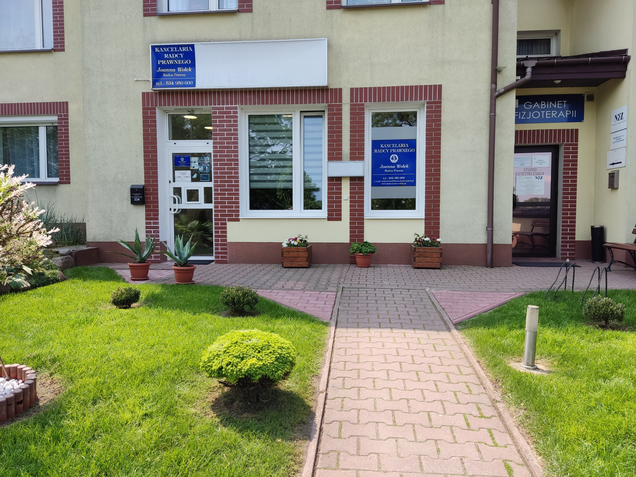 Wejście do budynku z tabliczkami informacyjnymi o Kancelarii Radcy Prawnego Joanna Wołek i Gabinecie Fizjoterapii, z zadbanym trawnikiem, krzewami i kwiatami w donicach.