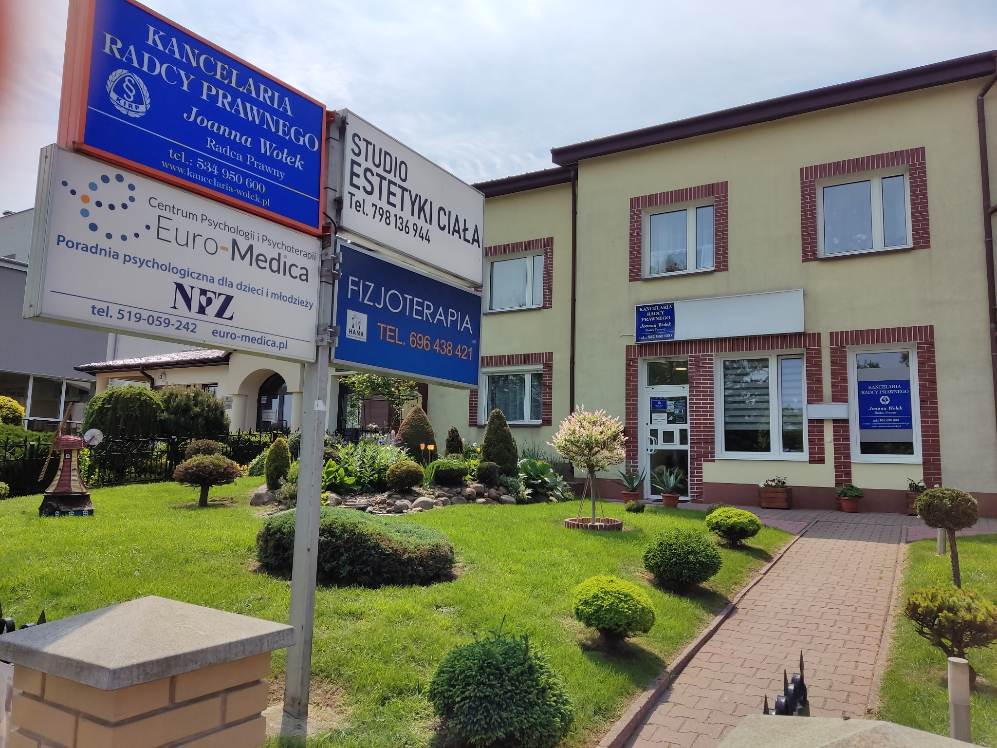 Budynek kancelarii radcy prawnego Joanna Wołek z tablicami informacyjnymi, zadbanym trawnikiem i ozdobnymi krzewami przed wejściem.