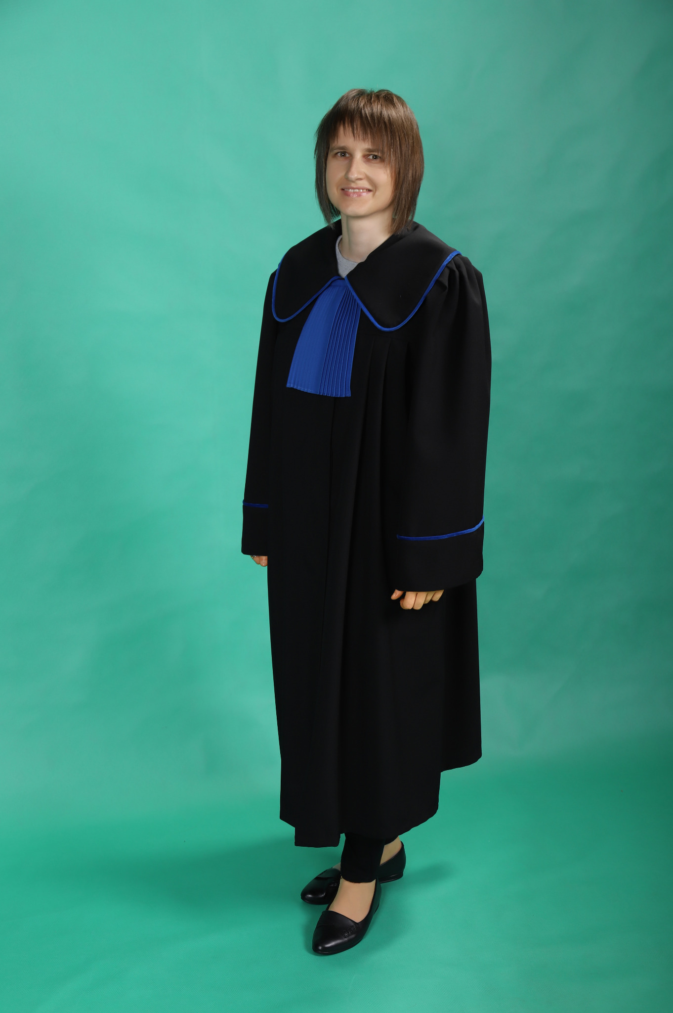 Uśmiechnięta kobieta w todze adwokackiej z niebieskimi plisami, stojąca na tle jednolitego, turkusowego tła.