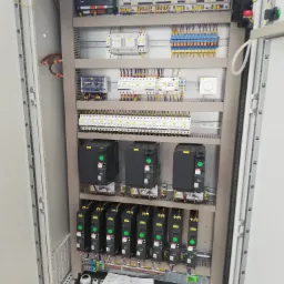 Otwarta szafa sterownicza z zainstalowanymi modułami elektronicznymi, zabezpieczeniami i okablowaniem, widoczne brązowe przewody u góry po lewej, dokumentacja techniczna na dole szafy.