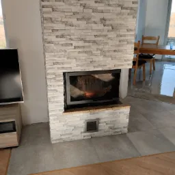 Nowoczesny kominek obłożony szarą, imitującą kamień okładziną, z widocznym paleniskiem i kratką wentylacyjną u dołu, na tle szarej podłogi i fragmentu drewnianej podłogi.