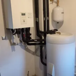 Biała pompa ciepła Panasonic Aquarea z czarnymi rurami instalacji grzewczej oraz zbiornikiem buforowym w pomieszczeniu technicznym, widoczne elementy instalacji wodnej z zaworami i manometrem.