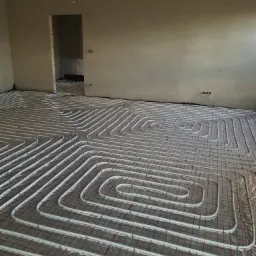 Białe rurki ogrzewania podłogowego ułożone spiralnie na siatce w surowym wnętrzu, widoczne niedokończone ściany i otwór drzwiowy.