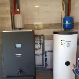 Kotłownia z kotłem na pellet Kostrzewa, zbiornikiem ciepłej wody użytkowej Kospel i instalacją hydrauliczną w pomieszczeniu wyłożonym płytkami.