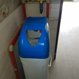 Niebiesko-biały zmiękczacz wody podłączony do instalacji z czerwonych rur, z filtrem mechanicznym i regulatorem ciśnienia, na tle jasnych płytek ceramicznych.