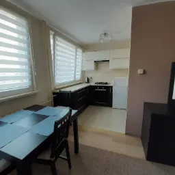 Widok na odnowione wnętrze mieszkania z aneksem kuchennym, widoczne żaluzje okienne, stół z krzesłami i telewizor na komodzie.