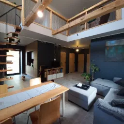 Wnętrze domu z drewnianymi schodami spiralnymi, drewnianym stołem jadalnym z szarą bieżnią, szarą sofą narożną i antresolą z drewnianymi belkami oraz siatką zabezpieczającą.