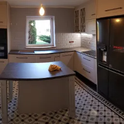 Jasna kuchnia z wyspą, czarnym blatem i wzorzystą podłogą. W tle okno z widokiem na zieleń i czarna lodówka LG.