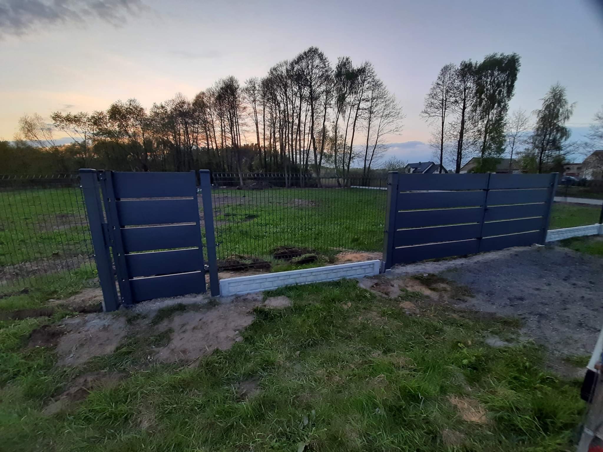 Nowoczesne, grafitowe ogrodzenie panelowe z poziomymi listwami i siatką, widoczne na tle zielonego trawnika i drzew o zachodzie słońca.