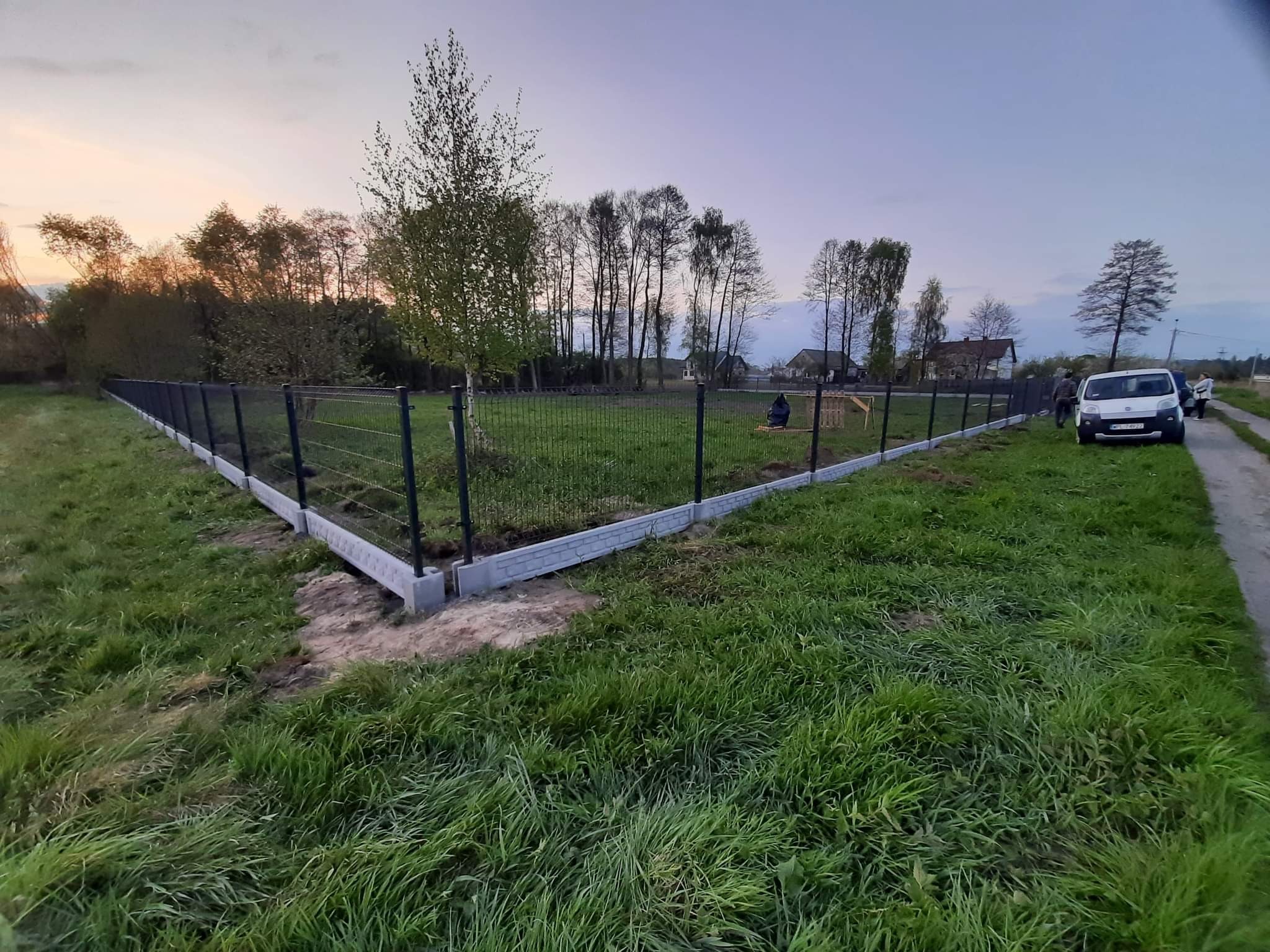 Nowo postawione ogrodzenie panelowe z podmurówką z bloczków betonowych, widok na posesję i okolicę w tle o zmierzchu.