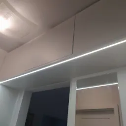 Białe szafki pod sufitem z oświetleniem LED, minimalistyczny design, widoczne lustro i wieszak na ścianie.