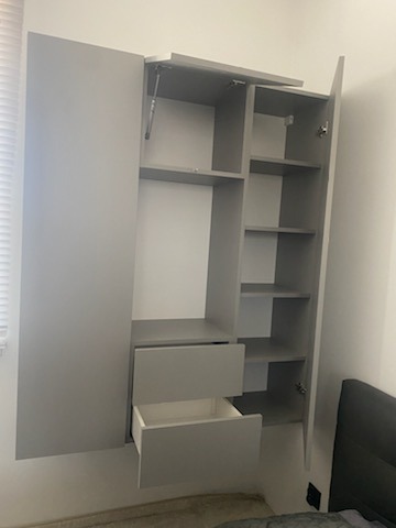 Szara szafa z otwartymi drzwiami i szufladami, widoczne półki, zamontowana na ścianie w pomieszczeniu z oknem i łóżkiem.