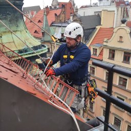 BEST TEAM s.c - Pracownik w kasku, uprzęży i rękawicach, zabezpieczony linami, czyści metalową konstrukcję na dachu zabytkowego budynku z czerwonej dachówki; widoczne inne dachy w tle.