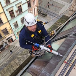 BEST TEAM s.c - Pracownik w kasku, zabezpieczony linami, uszczelnia silikonem obróbkę blacharską dachu zabytkowej kamienicy, widok z góry na ulicę z przechodniami.