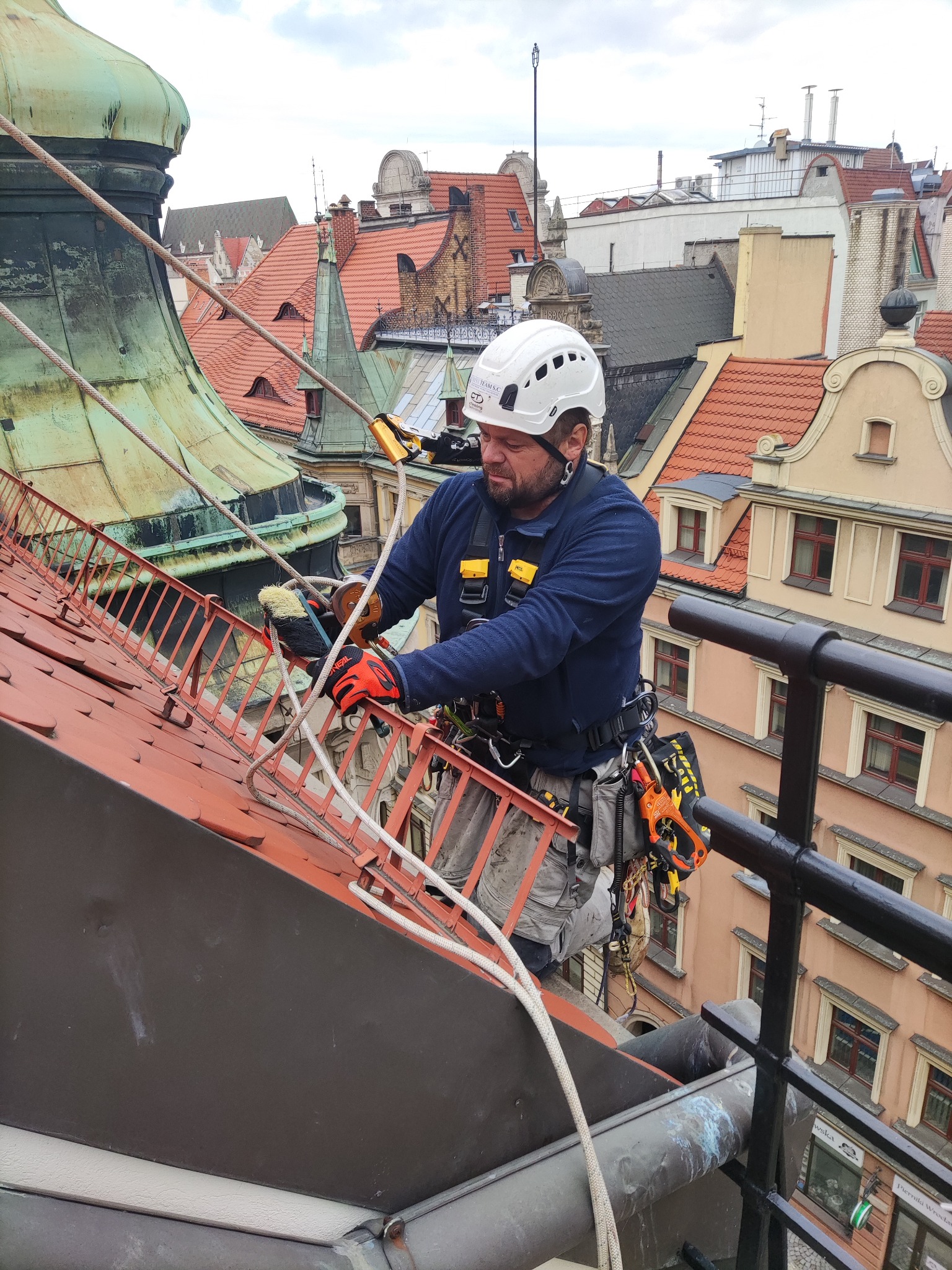Pracownik w kasku, uprzęży i rękawicach, zabezpieczony linami, czyści metalową konstrukcję na dachu zabytkowego budynku z czerwonej dachówki; widoczne inne dachy w tle.