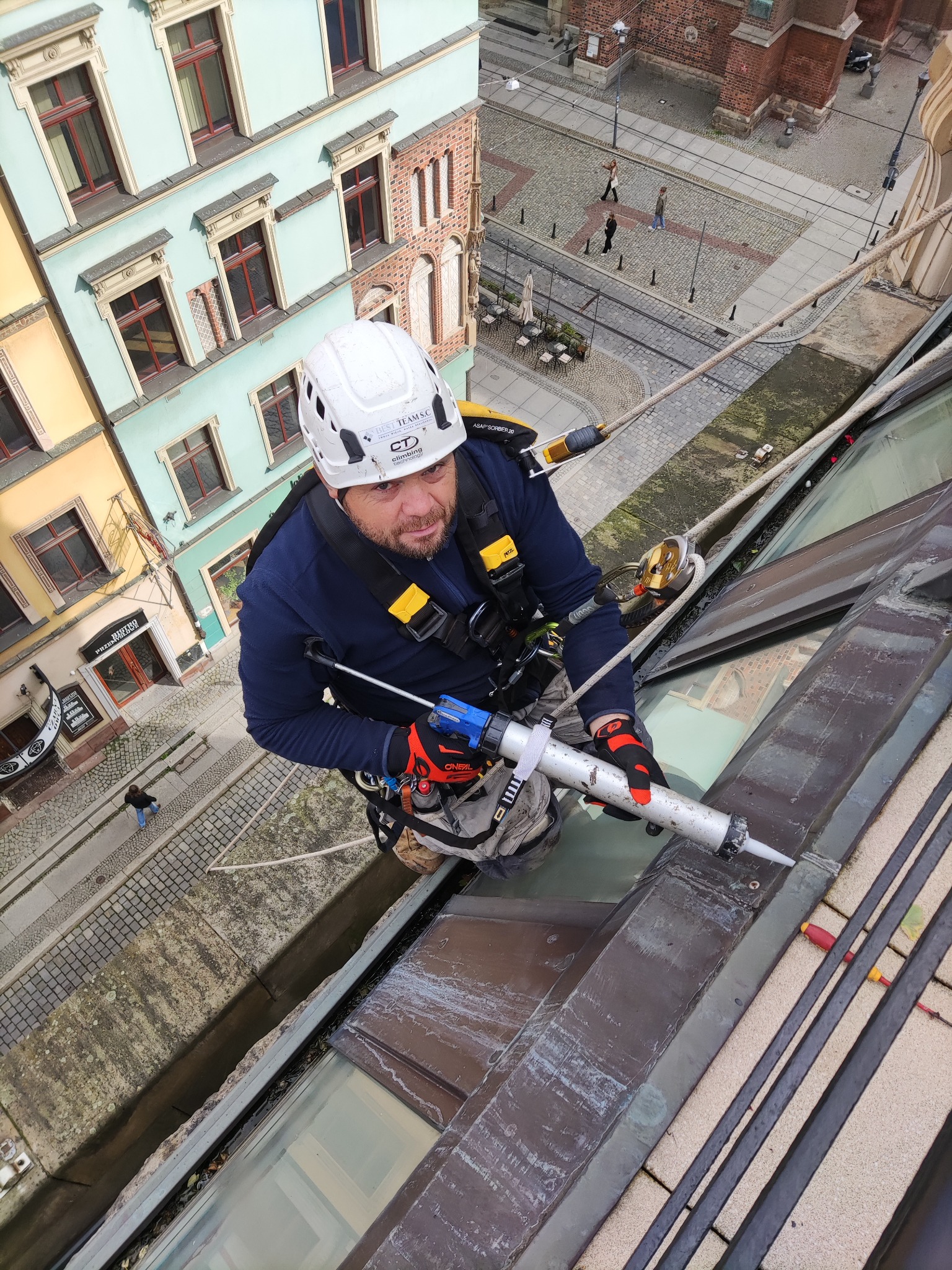 Pracownik w kasku, zabezpieczony linami, uszczelnia silikonem obróbkę blacharską dachu zabytkowej kamienicy, widok z góry na ulicę z przechodniami.