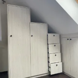 Biała szafa wnękowa z drewnopodobnym dekorem, zamontowana na poddaszu ze skosami, z otwartymi szufladami z czarnymi gałkami.