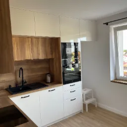 Nowoczesna kuchnia z białymi i drewnianymi frontami, czarną armaturą, piekarnikiem Electrolux i widokiem na osiedle przez okno.